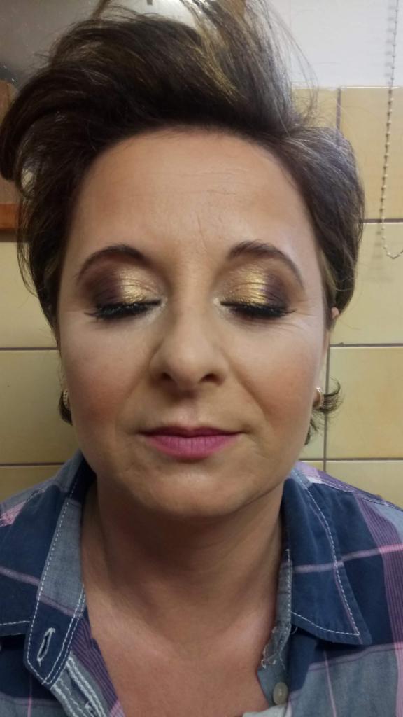 Kobieta z zamkniętymi oczami prezentuje złoty makijaż oczu i różowe usta, ubrana w koszulę w kratę, widoczne kolczyki.