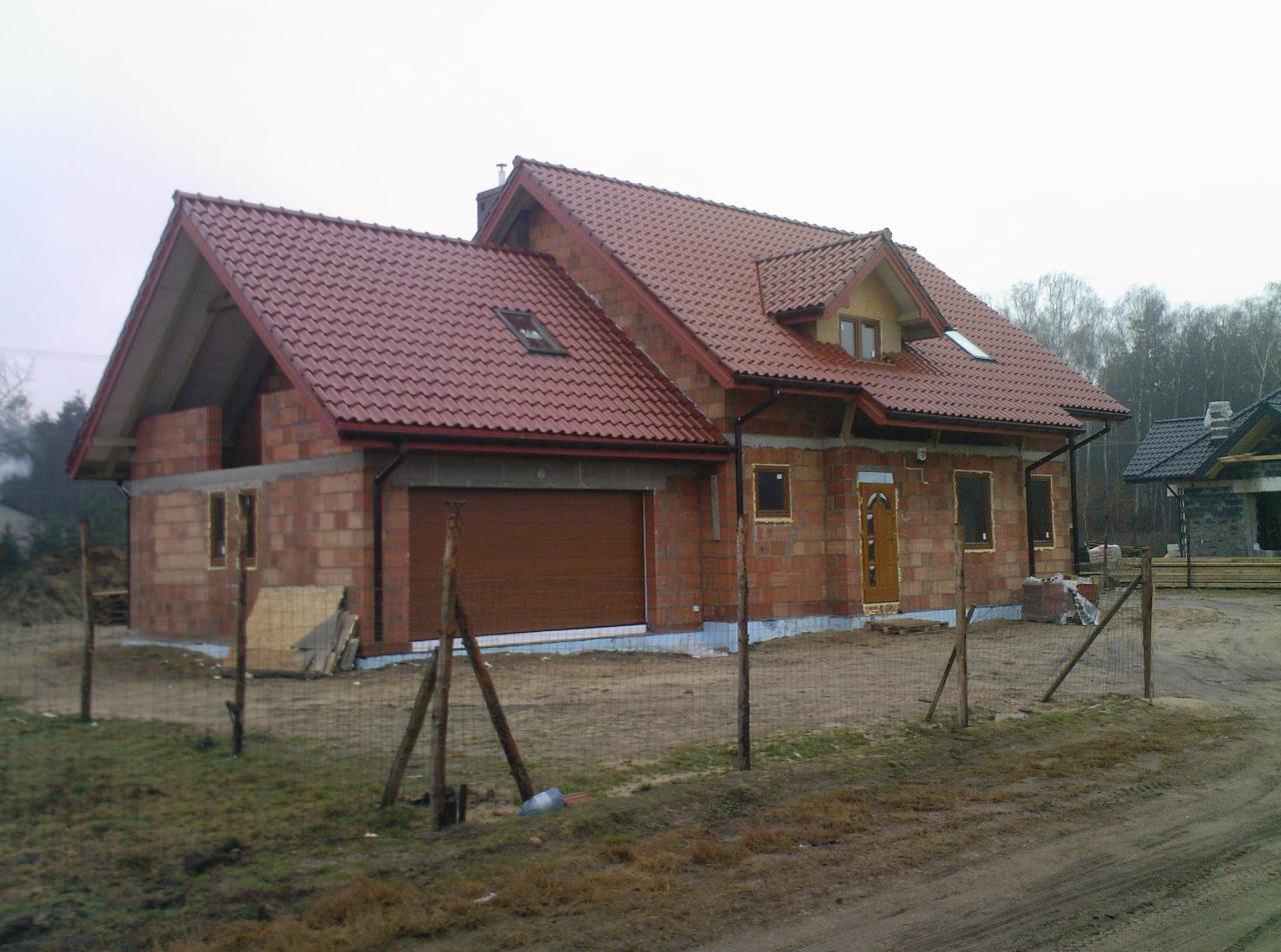 Budowa domu jednorodzinnego w stanie surowym otwartym, widoczne ściany z cegły, czerwona dachówka, brama garażowa, teren ogrodzony siatką.
