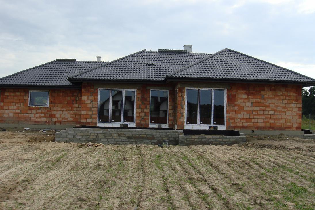 Surowy budynek jednorodzinny w trakcie budowy, widoczne ceglane ściany, szara dachówka, zamontowane okna, teren wokół budynku nierówny, piaszczysty.