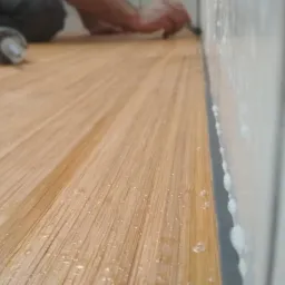 Szczegółowe ujęcie układania paneli podłogowych, z widocznym klejem na ścianie i narzędziami w tle, ukazujące precyzję wykończenia.