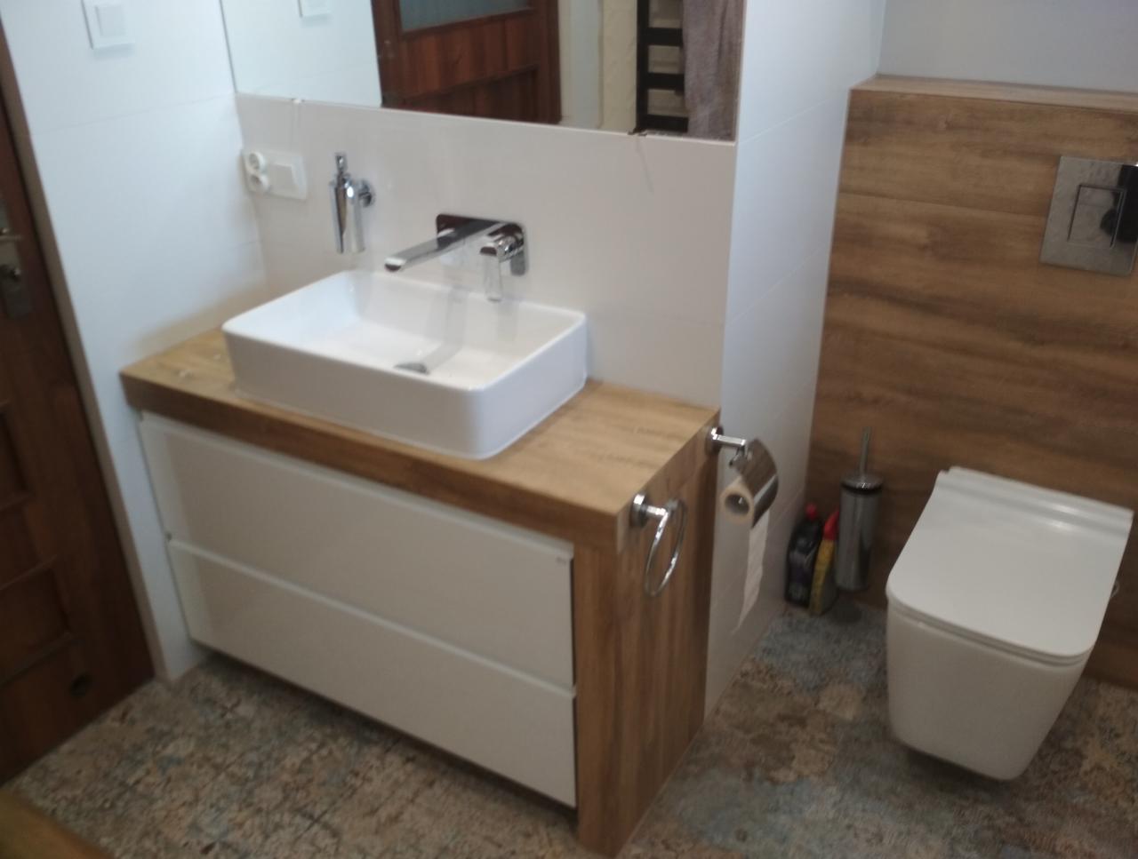 Nowoczesna łazienka z wiszącą toaletą, prostokątną umywalką nablatową na szafce z drewnianym blatem i białymi frontami, oraz drewnopodobną okładziną ścienną.