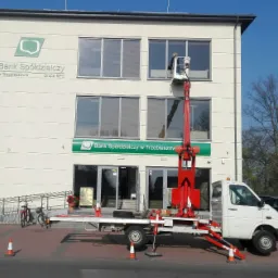 Pracownik na wysięgniku myje okna Banku Spółdzielczego w Trzebieszowie, widoczny biały budynek banku, czerwony podnośnik koszowy i zaparkowane samochody.