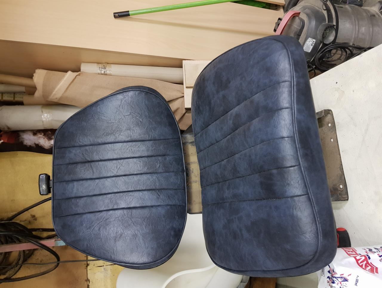 Dwa odnowione, granatowe siedziska z poziomymi przeszyciami, jedno z metalowym mocowaniem, drugie z czarnym uchwytem, na tle warsztatu z materiałami i narzędziami.