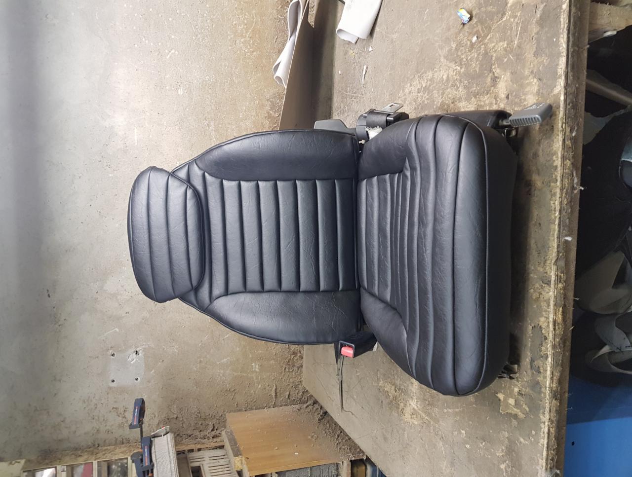Odnowione czarne skórzane siedzenie samochodowe z pionowymi przeszyciami, widoczny pas bezpieczeństwa, umieszczone na warsztatowym stole.
