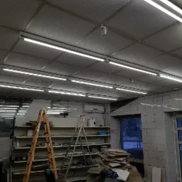 Wnętrze pomieszczenia w trakcie prac, widoczne drabiny, płytki na ścianach, zamontowane lampy jarzeniowe pod sufitem, regały z materiałami i kartonami.