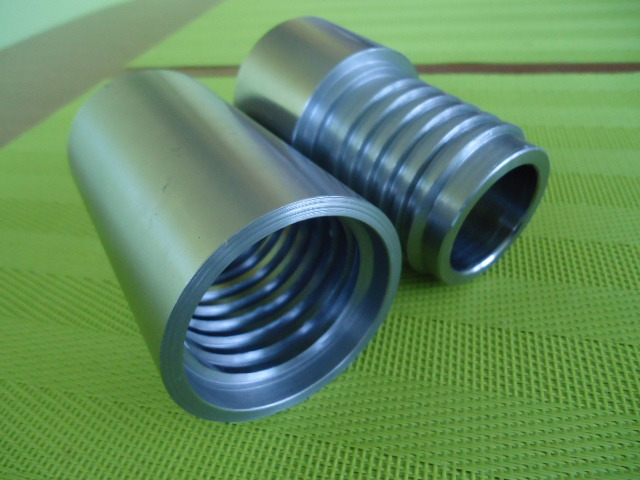 Dwa metalowe cylindryczne elementy, jeden gładki, drugi z rowkami, leżące na zielonej, teksturowanej powierzchni, widoczne ślady obróbki.