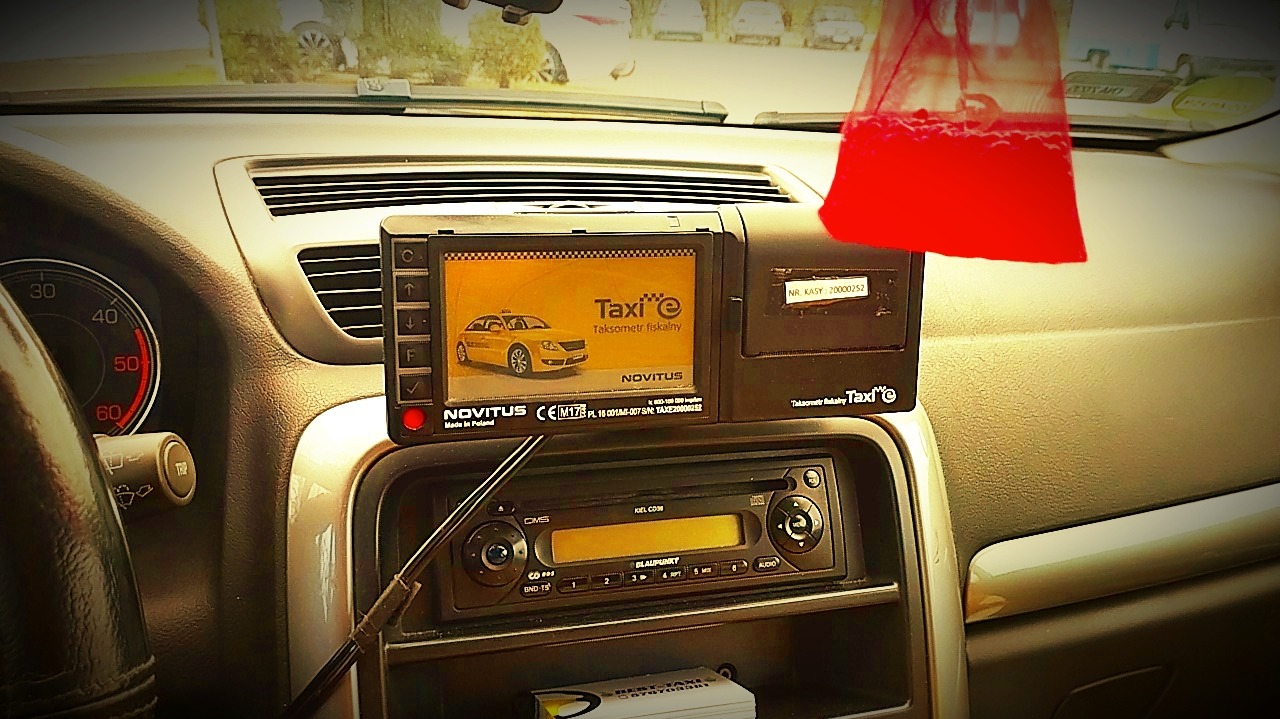 Wnętrze taksówki: taksometr fiskalny Novitus z żółtym autem na ekranie, radio Blaupunkt i czerwona zawieszka zapachowa. Widoczny fragment kierownicy i deski rozdzielczej.