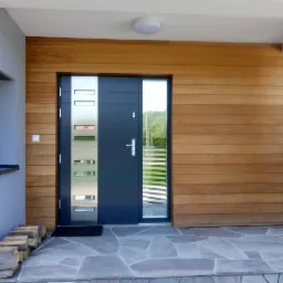 Nowoczesne wejście do domu z elewacją z jasnego drewna, szarą stolarką okienną i drzwiową z elementami metalowymi oraz kamienną posadzką.