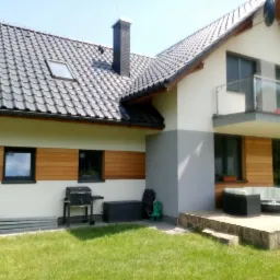 Nowoczesny dom z tarasem wyłożonym kamieniem, meble ogrodowe z białymi poduszkami, drewniane panele elewacyjne między oknami, zadbany trawnik.