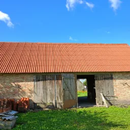Budynek gospodarczy z cegły z nową, czerwoną dachówką karpiówką, otwartymi drewnianymi wrotami i stertą dachówek obok.