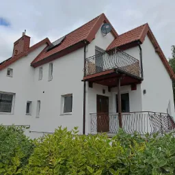 Biały dom z czerwonym dachem i balkonami z ozdobnymi, brązowymi balustradami. Widoczne panele słoneczne na dachu i antena satelitarna. Krzewy w pierwszym planie.