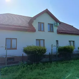 Biały dom z brązowym dachem i oknami, otoczony zielenią. Widoczny klimatyzator na ścianie. Metalowe ogrodzenie i krzewy przed budynkiem. Słoneczny dzień.