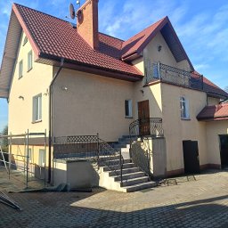 Putz-Mal - Dwupiętrowy dom z beżową elewacją, czerwonym dachem i balkonem z kutą balustradą. Widoczne schody wejściowe, okna i elementy wykończeniowe. Błękitne niebo w tle.