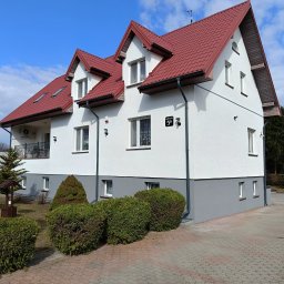 Putz-Mal - Dwukondygnacyjny dom z czerwonym dachem i jasną elewacją, widok z poziomu kostki brukowej. Widoczne rynny, okna i adres na budynku. Błękitne niebo z chmurami w tle.