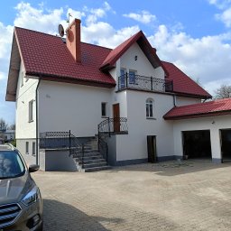 Putz-Mal - Biała elewacja domu z czerwonym dachem w Olecku, widok z zewnątrz. Schody z czarną balustradą prowadzą do drzwi wejściowych. Obok garaż z otwartymi wrotami.