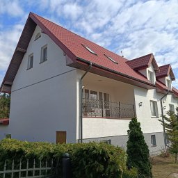 Putz-Mal - Dwupiętrowy dom z czerwonym dachem i balkonem, otoczony zielenią. Widoczne rynny i okna dachowe. Jasna elewacja kontrastuje z błękitnym niebem.