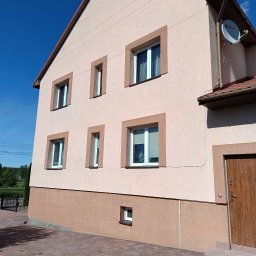 Putz-Mal - Dwukondygnacyjny dom z elewacją w odcieniach beżu, z brązowymi obramowaniami okien i dachem. Widoczna antena satelitarna i kostka brukowa przed budynkiem.