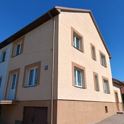 Putz-Mal - Dwukondygnacyjny dom z beżową elewacją i brązowymi akcentami, widoczny z perspektywy żabiej. Rynny w kolorze dachu. Numer domu '20' na ścianie. Błękitne niebo w tle.