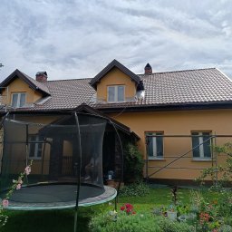 Putz-Mal - Dwupiętrowy dom z brązową dachówką i jasnopomarańczową elewacją, częściowo otoczony rusztowaniem. Na dachu widoczny pracownik, jedno okno na poddaszu zakryte folią. W ogrodzie trampolina i kwitnące...