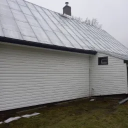 Boczny widok domu z metalowym dachem pokrytym lekkim śniegiem, białą elewacją z sidingu i widocznym kominem.