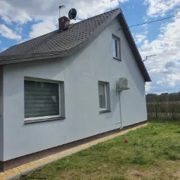 Szary dom jednorodzinny z brązowym dachem i widoczną instalacją klimatyzacyjną na elewacji bocznej, w tle błękitne niebo z chmurami.