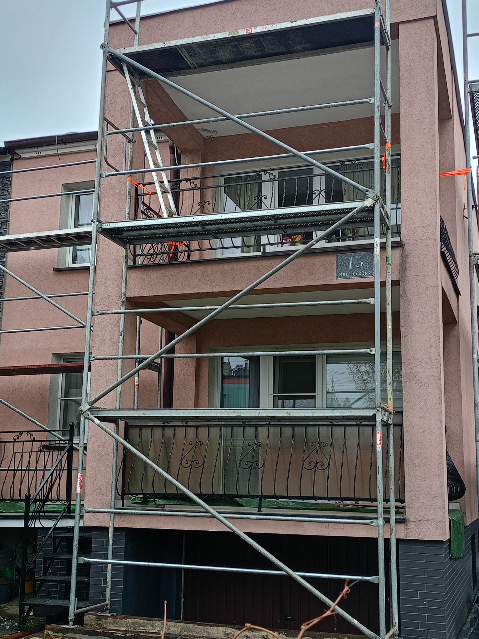Różowa elewacja budynku mieszkalnego w trakcie prac. Widoczne rusztowania, balkony z ozdobnymi balustradami i tabliczka z numerem domu. Detal elewacji.