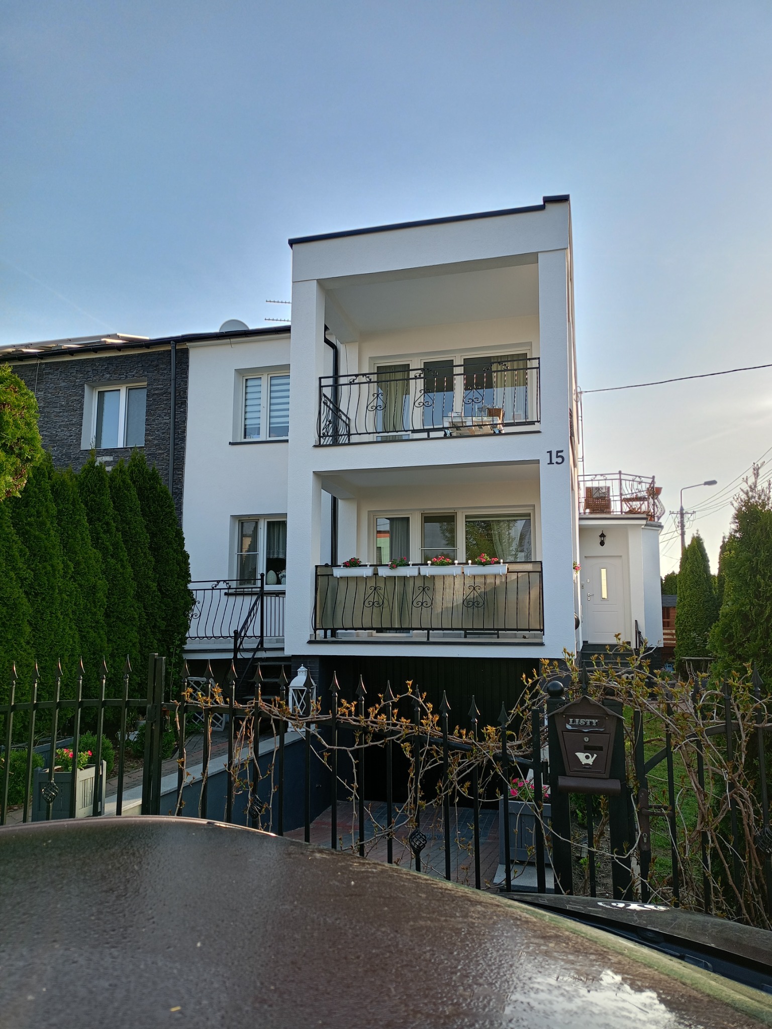 Biały, dwupiętrowy dom z balkonami o żeliwnych balustradach, numerem 15 na elewacji i skrzynką na listy przy ogrodzeniu. Widok z dołu, w tle błękitne niebo.