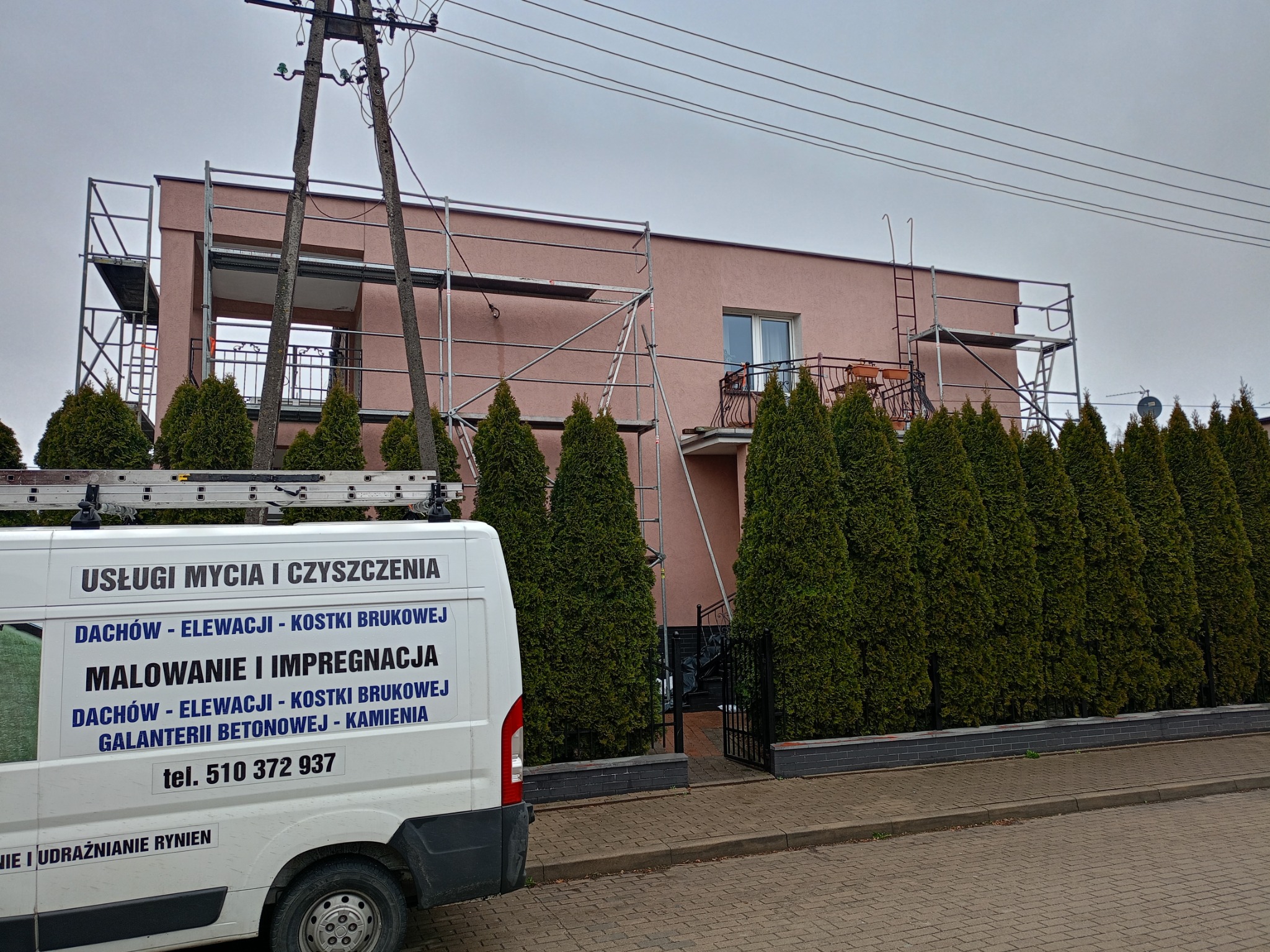 Różowa elewacja domu w rusztowaniach, widoczny fragment firmowego auta z ofertą mycia i malowania. Zielone tuje przed budynkiem.