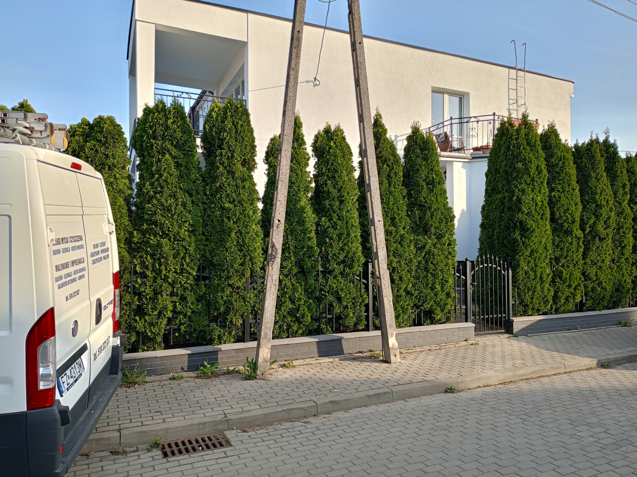 Biały bus z napisem o usługach mycia i malowania, stoi na tle nowoczesnego domu z balkonem i rzędem tuj. Widoczny słup energetyczny i metalowa brama.