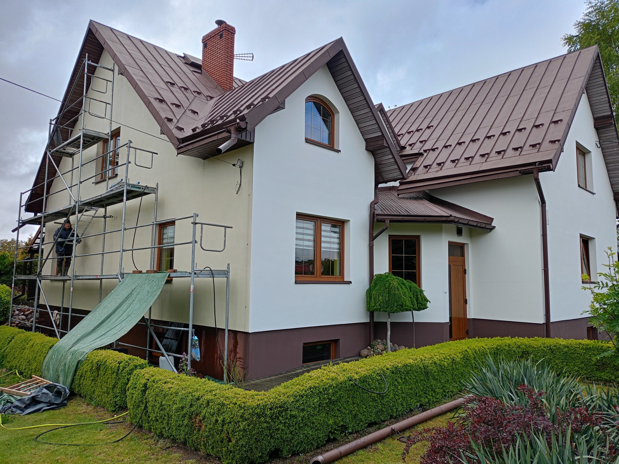 Dwukondygnacyjny dom z brązowym dachem i białą elewacją. Część ściany w kolorze żółtym, na rusztowaniu pracownik. Renowacja w toku.