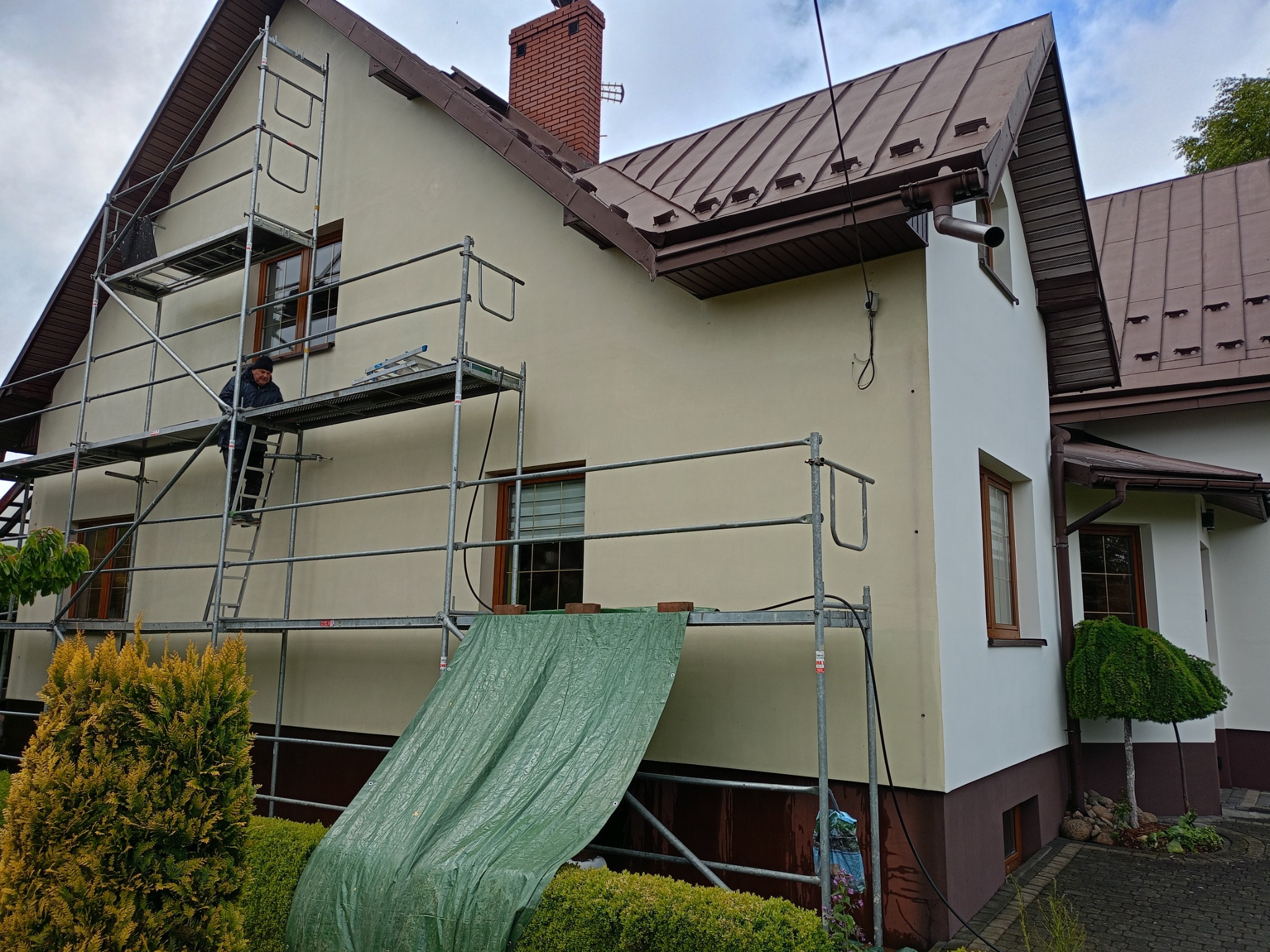 Pracownik na rusztowaniu maluje elewację domu. Widoczne zabezpieczenia, świeżo pomalowana część ściany na biało i beżowa elewacja. Ceglany komin i brązowy dach.