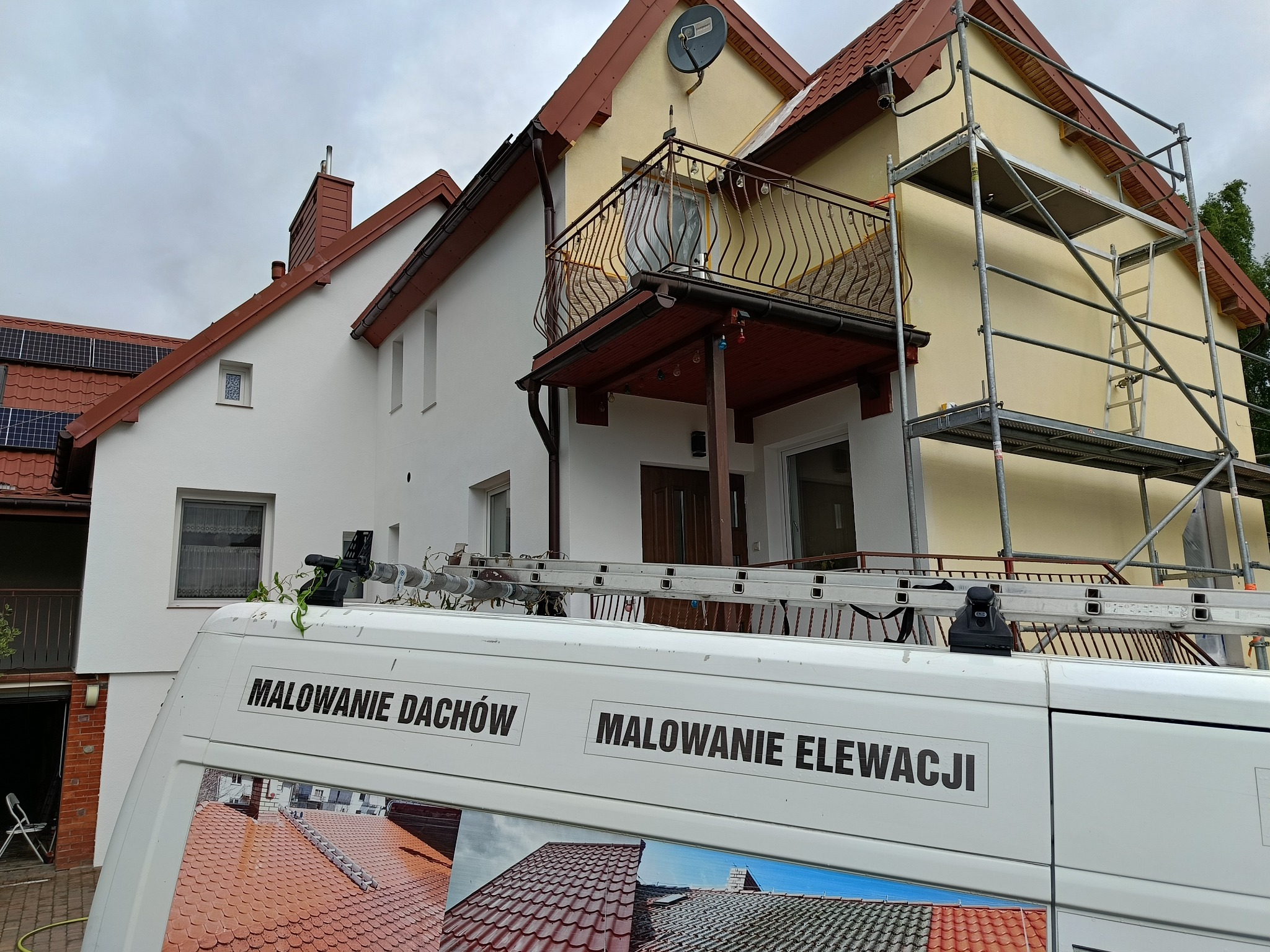 Dwukondygnacyjny dom z balkonem w trakcie malowania elewacji, widoczny zza samochodu firmowego z napisem 'Malowanie Dachów, Malowanie Elewacji' i rusztowaniem.