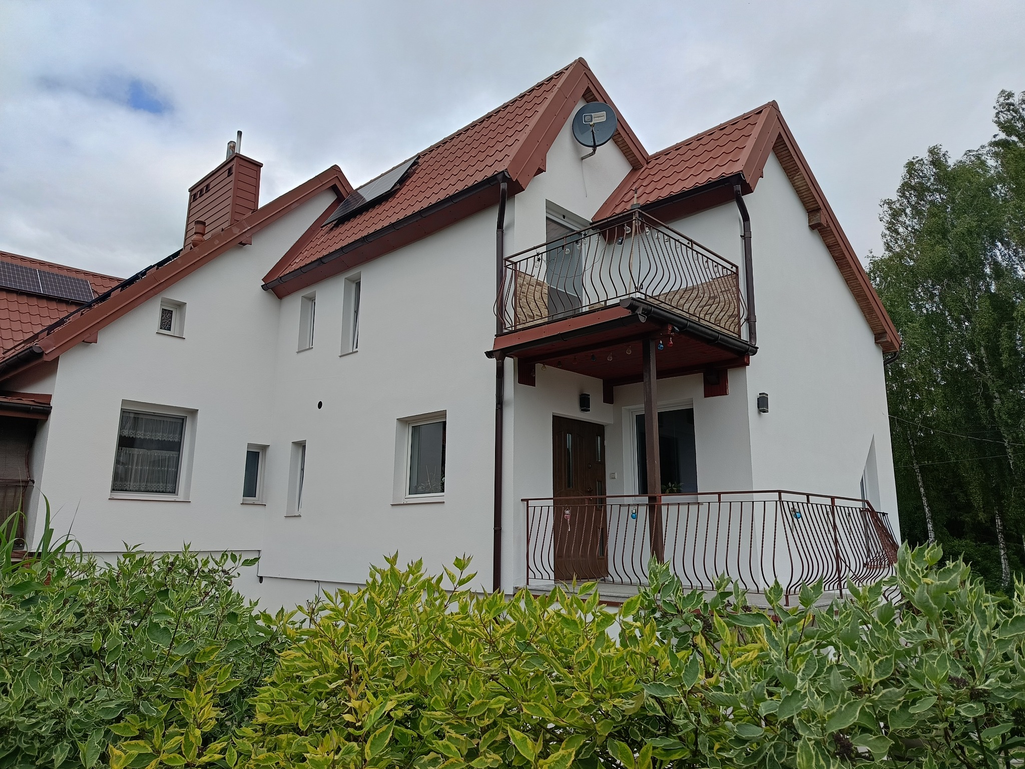 Biały dom z czerwonym dachem i balkonami z ozdobnymi, brązowymi balustradami. Widoczne panele słoneczne na dachu i antena satelitarna. Krzewy w pierwszym planie.