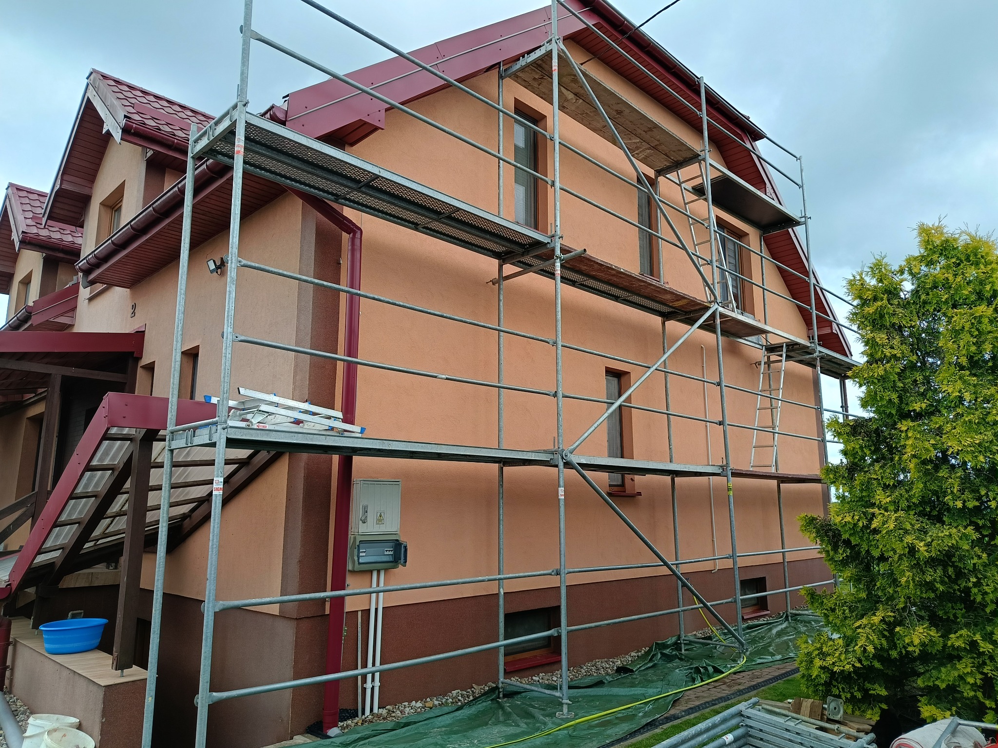 Dom jednorodzinny w trakcie prac elewacyjnych, osłonięty rusztowaniem. Widoczna pomalowana część fasady i przygotowane narzędzia.