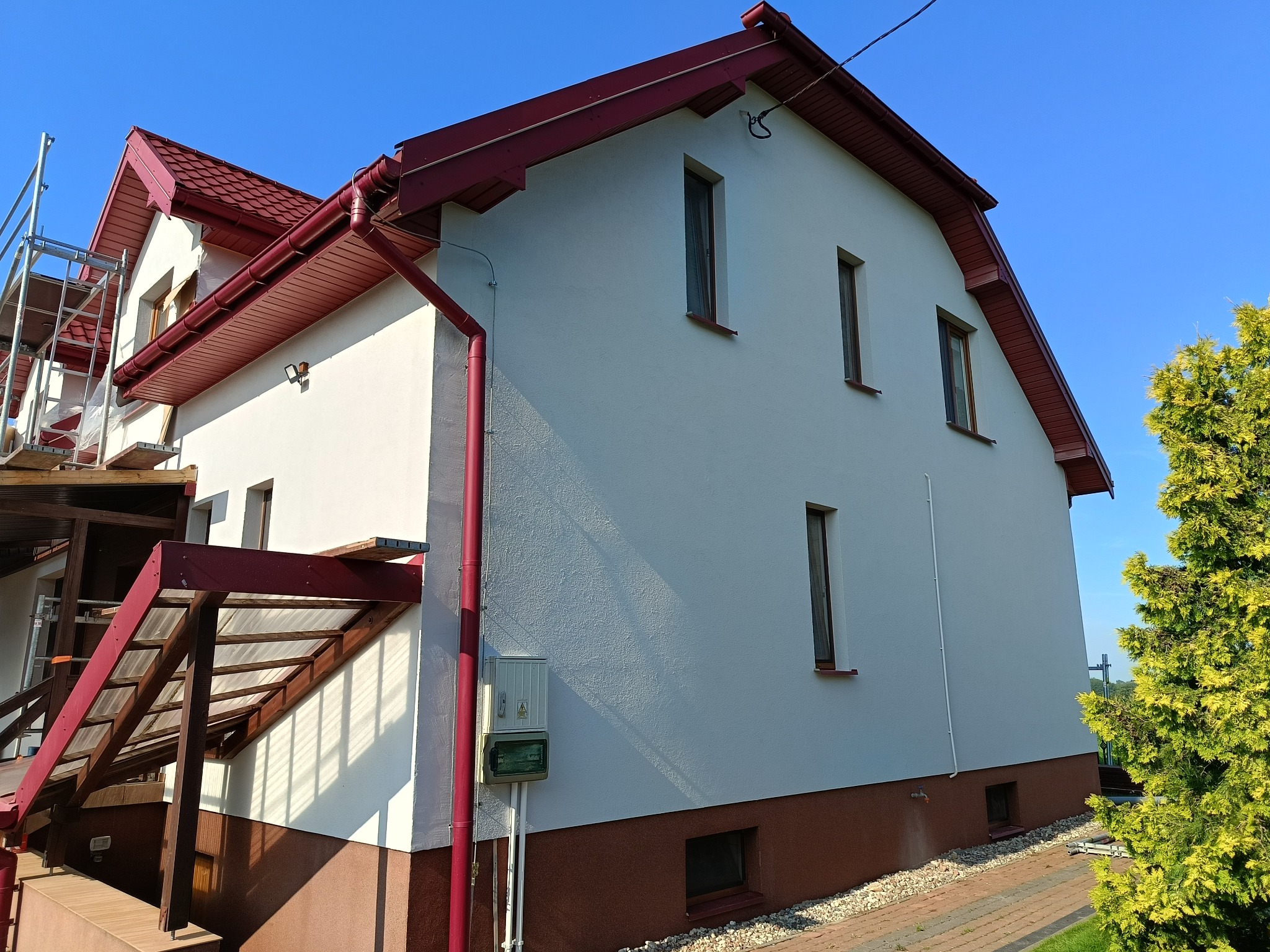 Biała elewacja domu z bordowym dachem i rynnami, widok z boku. Częściowo widoczne rusztowanie. Dolna część elewacji w kolorze brązowym.
