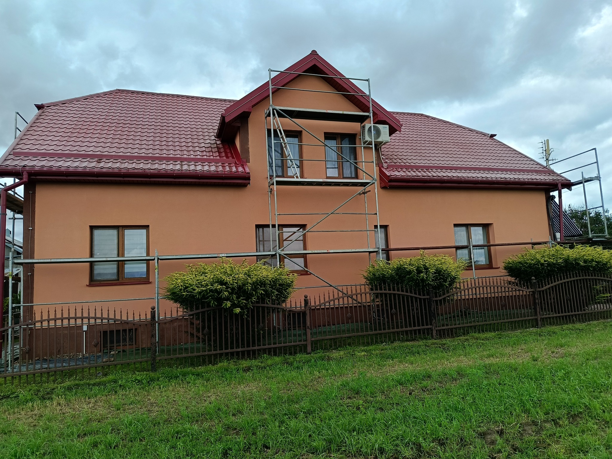 Dom z pomarańczową elewacją i czerwoną dachówką, częściowo osłonięty rusztowaniem. Widoczne okna z brązowymi ramami i zielony trawnik.