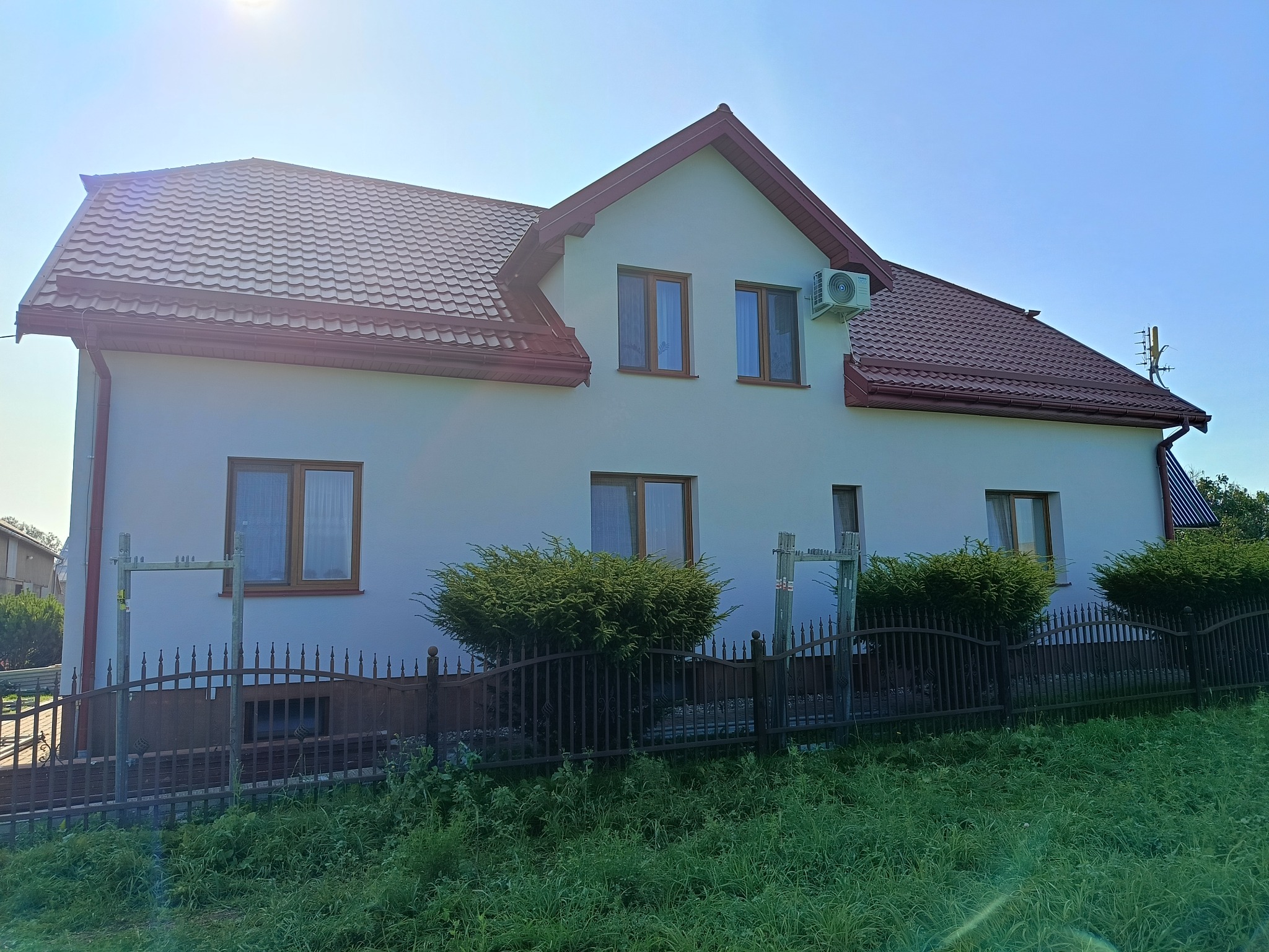 Biały dom z brązowym dachem i oknami, otoczony zielenią. Widoczny klimatyzator na ścianie. Metalowe ogrodzenie i krzewy przed budynkiem. Słoneczny dzień.