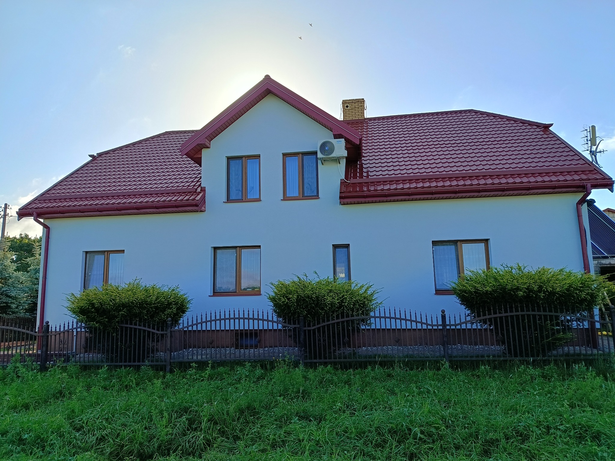 Dwupiętrowy dom z czerwoną dachówką, jasną elewacją, brązowymi ramami okiennymi i klimatyzacją na ścianie. Przed domem ozdobne krzewy i metalowe ogrodzenie.