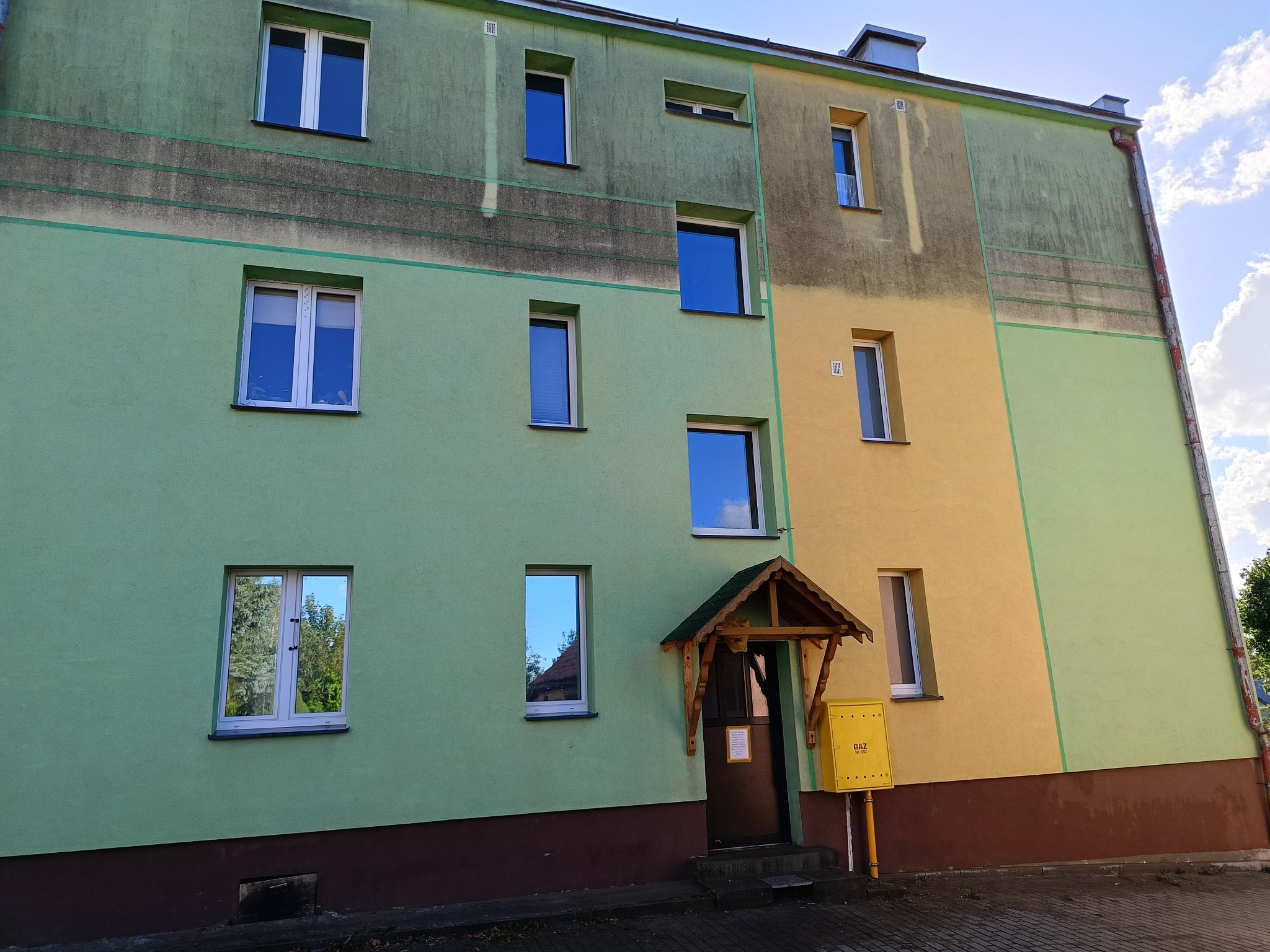 Wielopiętrowy budynek z elewacją w odcieniach zieleni i żółci, z widocznymi śladami zabrudzeń. Okna odbijają niebo i drzewa. Fragment chodnika i skrzynka gazowa.