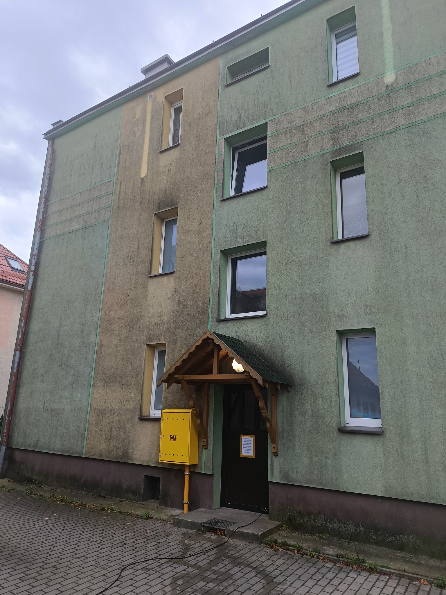Dwukolorowa elewacja budynku wielorodzinnego z widocznymi zabrudzeniami i zaciekami. Żółta skrzynka gazowa przy wejściu. Widoczne okna i zadaszenie nad drzwiami.