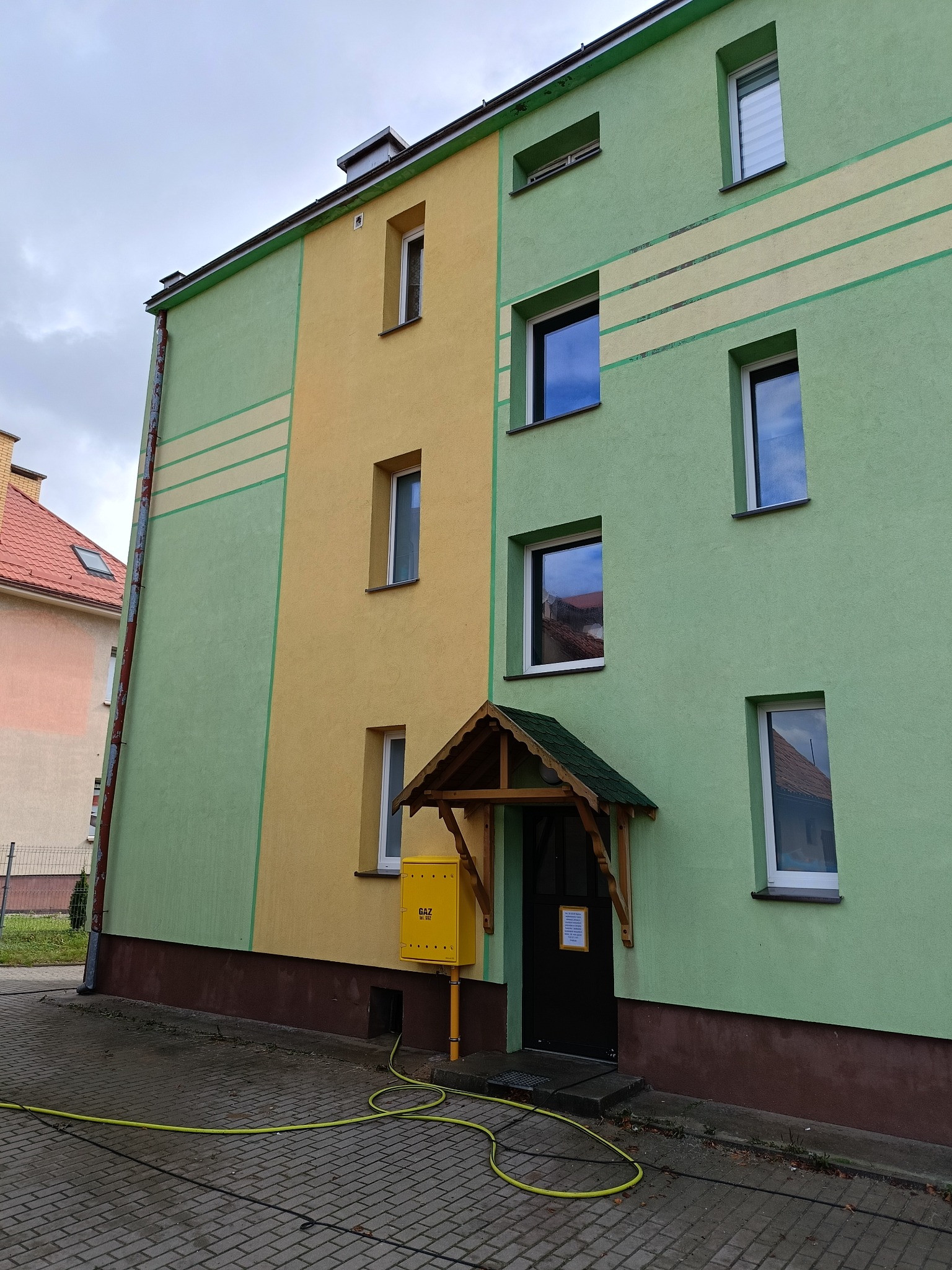 Odświeżona elewacja budynku wielorodzinnego w odcieniach zieleni i żółci. Widoczne linie dekoracyjne i żółta skrzynka gazowa przy wejściu.