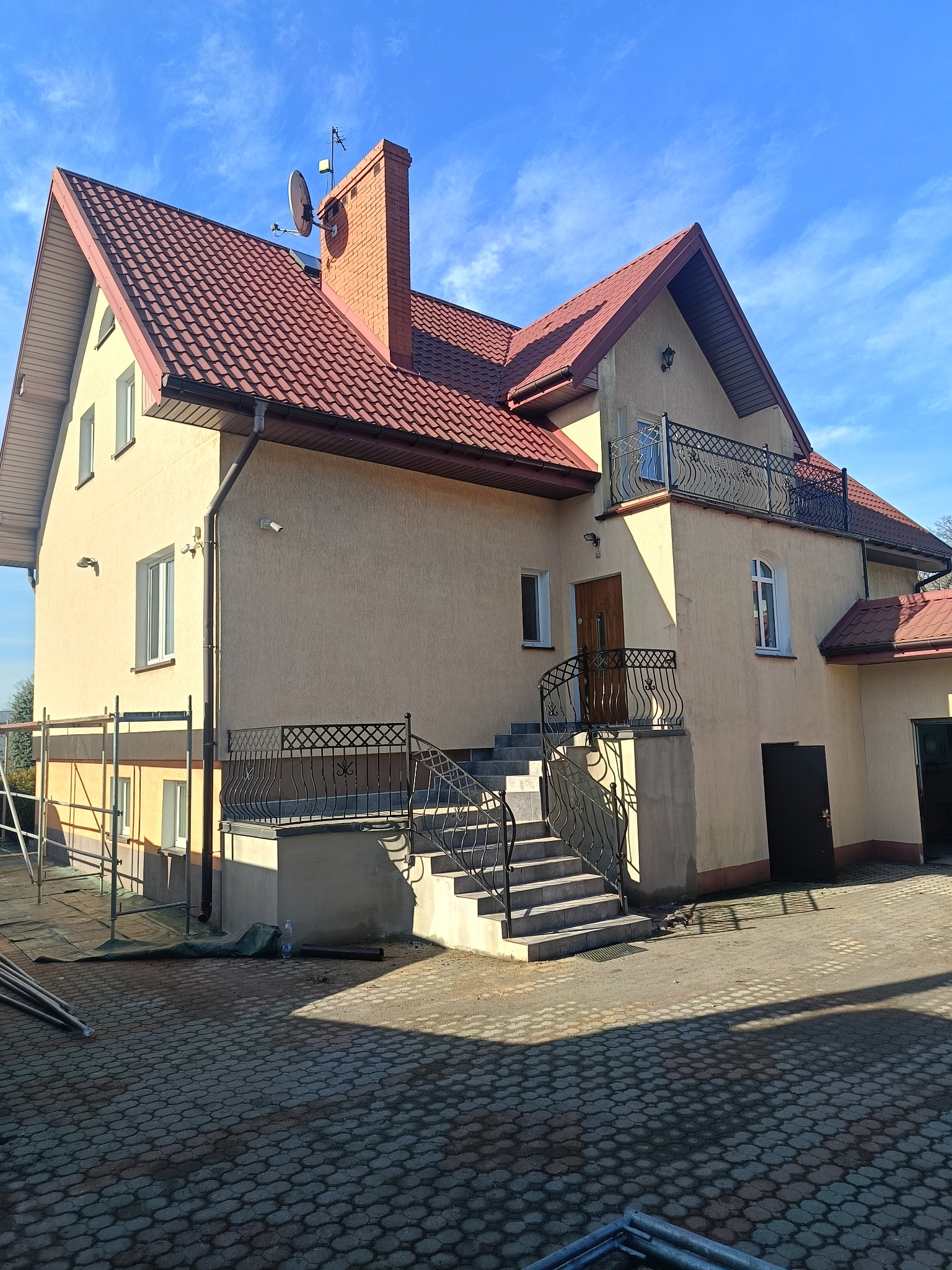 Dwupiętrowy dom z beżową elewacją, czerwonym dachem i balkonem z kutą balustradą. Widoczne schody wejściowe, okna i elementy wykończeniowe. Błękitne niebo w tle.