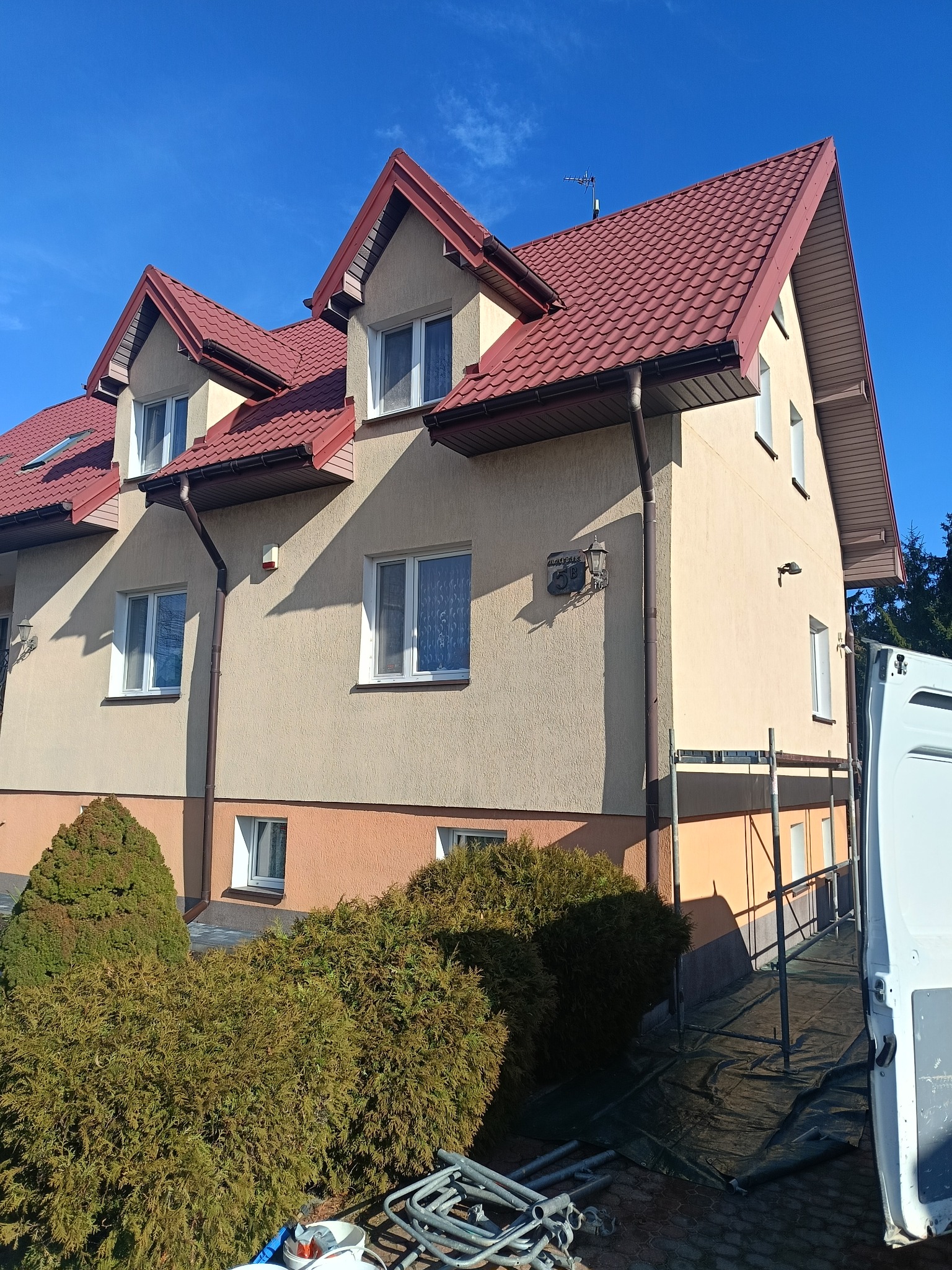 Dwupiętrowy dom z beżową elewacją i czerwonym dachem w słoneczny dzień. Widoczne rusztowania i częściowo samochód dostawczy. Ujęcie z dołu.