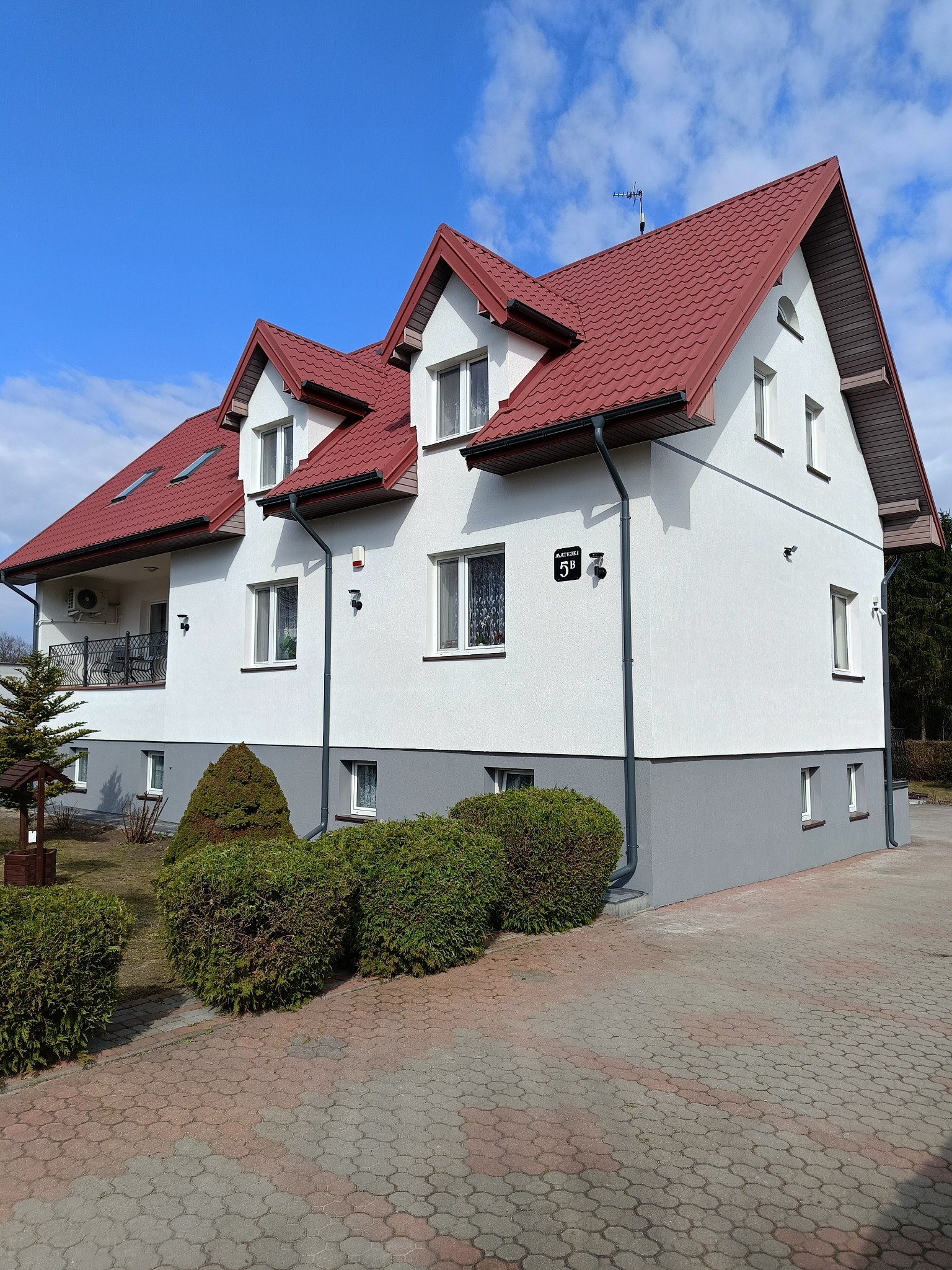 Dwukondygnacyjny dom z czerwonym dachem i jasną elewacją, widok z poziomu kostki brukowej. Widoczne rynny, okna i adres na budynku. Błękitne niebo z chmurami w tle.