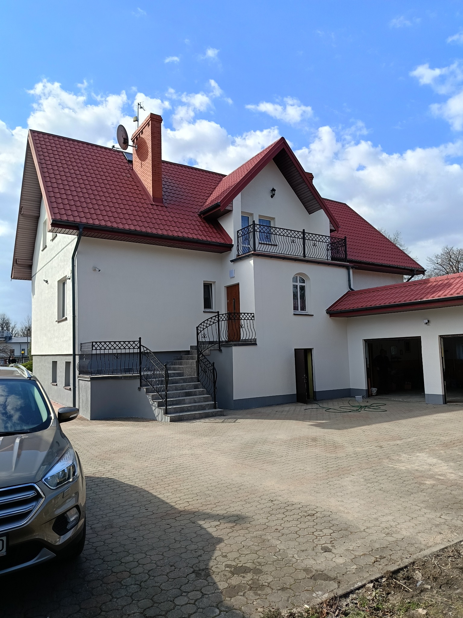 Biała elewacja domu z czerwonym dachem w Olecku, widok z zewnątrz. Schody z czarną balustradą prowadzą do drzwi wejściowych. Obok garaż z otwartymi wrotami.