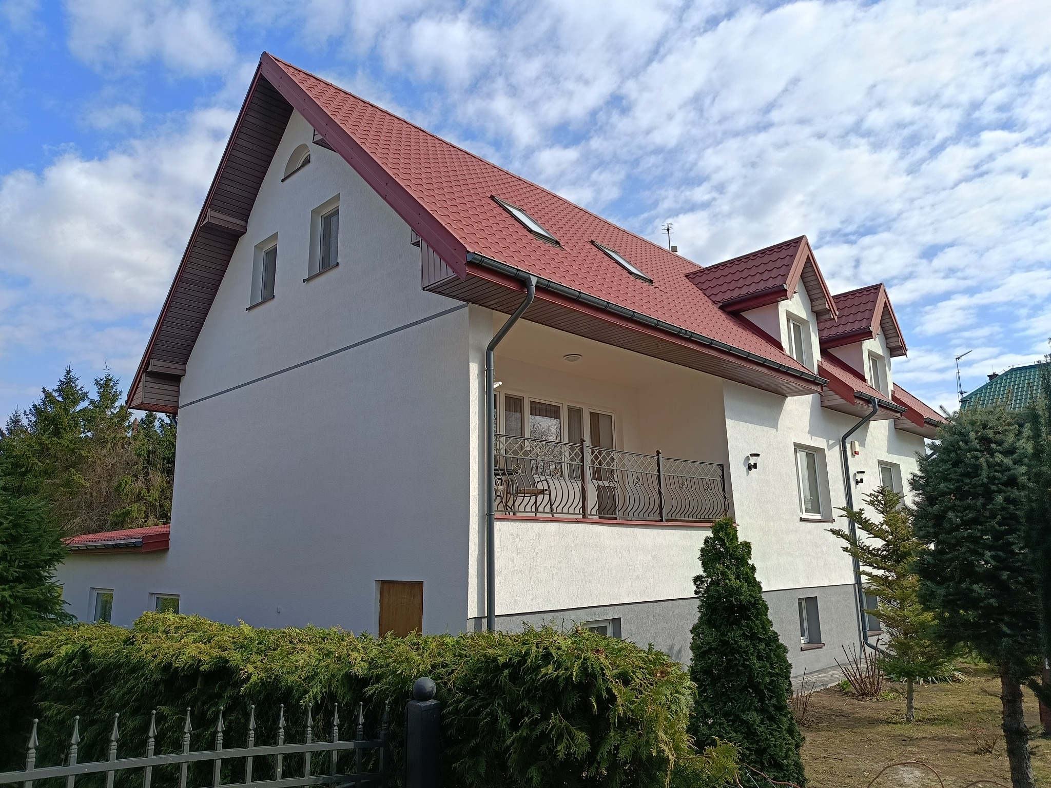 Dwupiętrowy dom z czerwonym dachem i balkonem, otoczony zielenią. Widoczne rynny i okna dachowe. Jasna elewacja kontrastuje z błękitnym niebem.