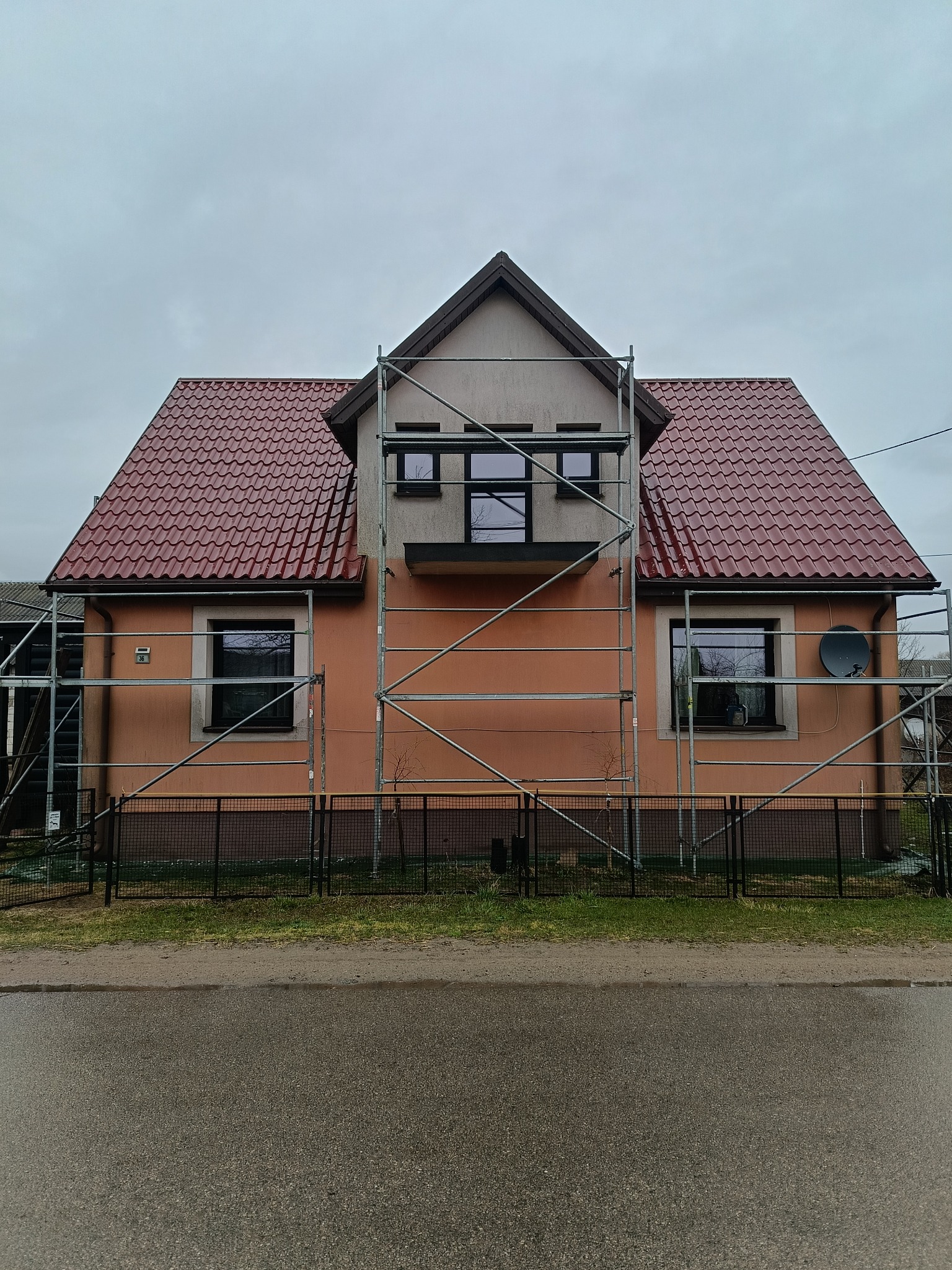 Dom jednorodzinny w trakcie malowania elewacji, otoczony rusztowaniem. Widoczny fragment dachu z czerwonej dachówki i szara, pochmurna pogoda.