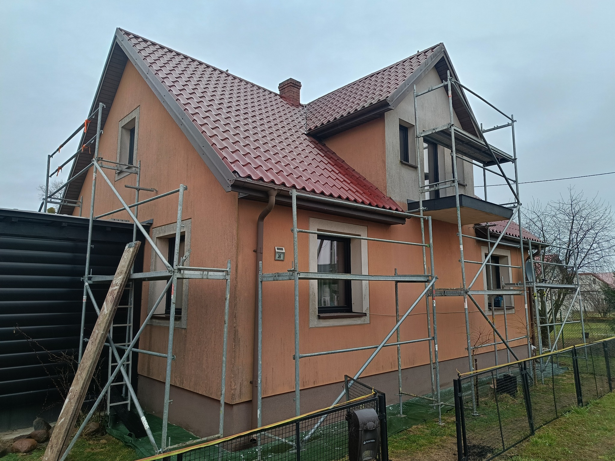 Dom jednorodzinny w rusztowaniach, z częściowo odnowioną elewacją i nowym, błyszczącym dachem z blachodachówki. Widoczny fragment numeru domu na elewacji.