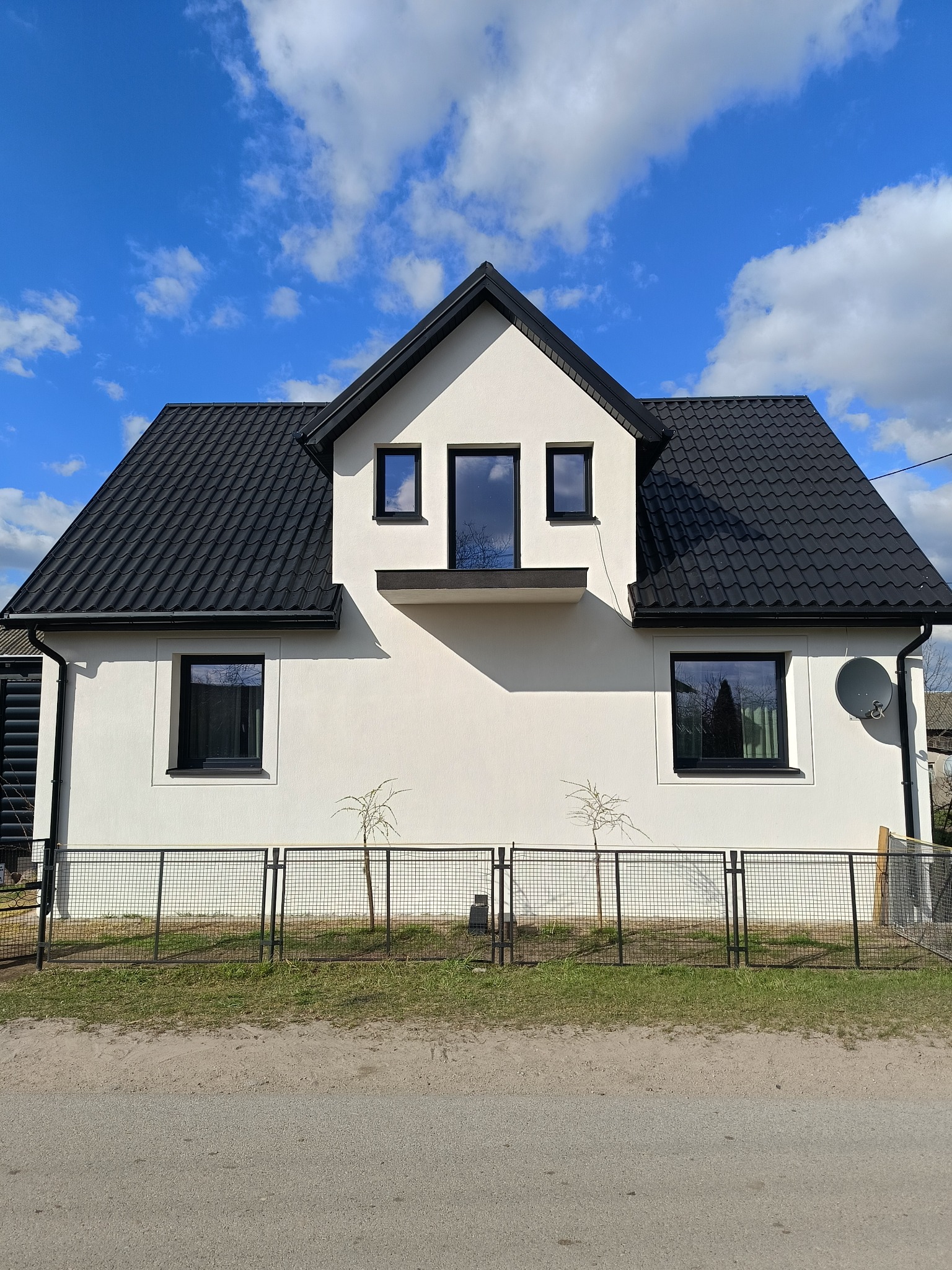 Biały dom z czarnym dachem i oknami, widziany z przodu, z metalowym ogrodzeniem i młodymi drzewkami. Błękitne niebo z chmurami w tle.