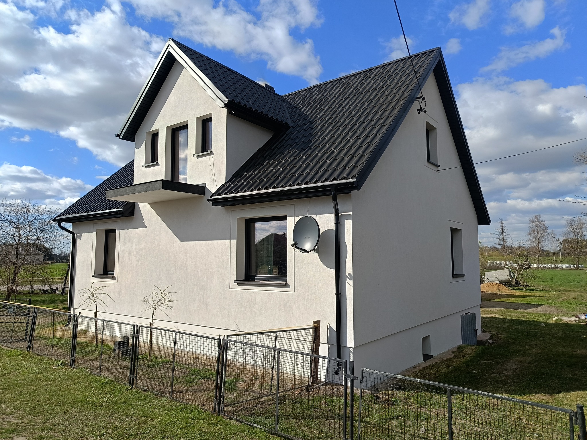 Dwukondygnacyjny dom z czarnym dachem i jasną elewacją, widziany z perspektywy ogrodu. Widoczne okna z czarnymi ramami i satelita na ścianie. Błękitne niebo z chmurami w tle.